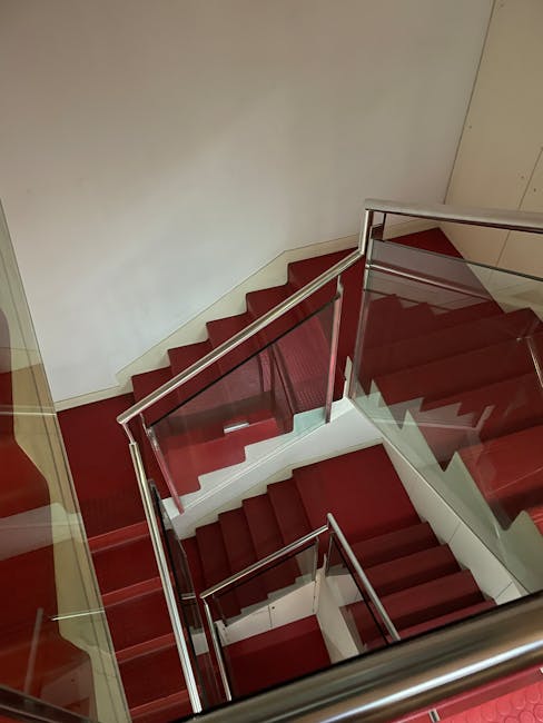 An aerial view of a modern stairwell with red carpeted steps, bordered by clear glass panels and stainless steel handrails. The walls are painted white, and the lighting is bright, illuminating the clean and well-maintained surfaces. The red carpet appears neatly laid, with no visible dust or stains, indicating recent cleaning or regular maintenance. The glass panels are transparent and spotless, contributing to an open and hygienic appearance. This space exemplifies effective surface cleaning and sanitisation practices in a residential or commercial building, highlighting the importance of maintaining hygiene standards for stairwell access, in line with Lambeth Cleaners' professional cleaning services. The image underscores the importance of careful attention to detail in cleaning high-traffic areas like stairwells within Lambeth estates, ensuring safety and cleanliness for residents and visitors alike.