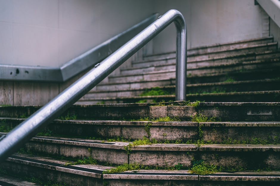 Close-up view of an outdoor concrete stairwell with moss and dirt accumulating between the steps, featuring a metallic handrail running diagonally across the image. The stairs show signs of weathering and grime, with some green moss and algae growth visible on the surface, indicating a need for deep cleaning and sanitisation. The area appears dimly lit, and the photo effectively captures the grime accumulation typical of outdoor stairwells in residential or commercial estates. This image illustrates the type of surfaces that Lambeth Cleaners can help maintain through professional surface cleaning and maintenance services, addressing access issues associated with moss and dirt buildup in stairwell environments.