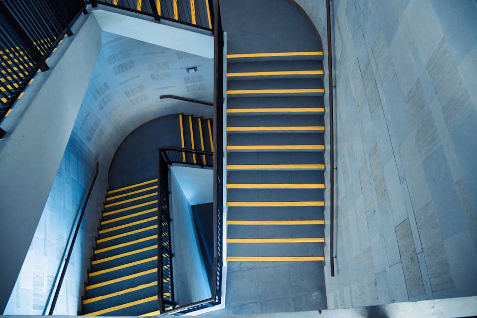 An aerial view of a modern stairwell with red carpeted steps, bordered by clear glass panels and stainless steel handrails. The walls are painted white, and the lighting is bright, illuminating the clean and well-maintained surfaces. The red carpet appears neatly laid, with no visible dust or stains, indicating recent cleaning or regular maintenance. The glass panels are transparent and spotless, contributing to an open and hygienic appearance. This space exemplifies effective surface cleaning and sanitisation practices in a residential or commercial building, highlighting the importance of maintaining hygiene standards for stairwell access, in line with Lambeth Cleaners' professional cleaning services. The image underscores the importance of careful attention to detail in cleaning high-traffic areas like stairwells within Lambeth estates, ensuring safety and cleanliness for residents and visitors alike.