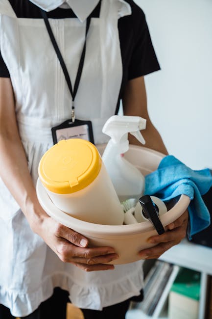 A person wearing a white and black uniform holds a round white cleaning caddy containing a yellow spray bottle, a white spray bottle, a blue microfiber cloth, and a black scrub brush, positioned in front of a plain white wall. The background includes part of a desk or shelf with some green and white folders or books. This image depicts tools used in professional domestic cleaning, focusing on surface cleaning and sanitisation, relevant to the services offered by Lambeth Cleaners for end of tenancy cleaning in Brixton SW2.