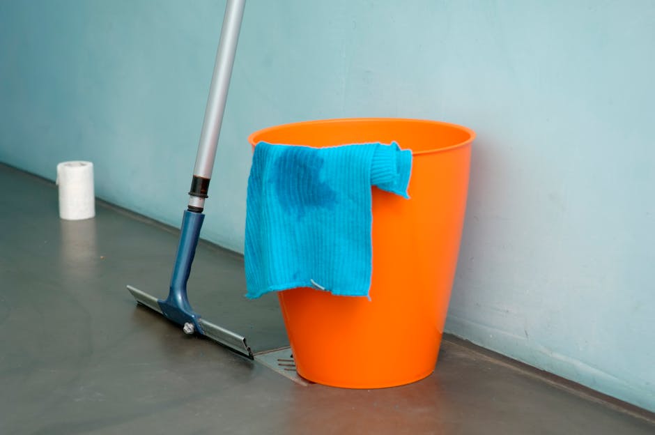 An orange plastic bucket with a blue microfiber cloth draped over its rim is positioned on a smooth gray floor next to a silver mop with a blue handle, which is resting against the bucket. Behind the bucket, a roll of white paper towels is visible near a light blue wall. The floor appears clean, and the scene suggests surface cleaning or maintenance in a commercial or residential setting, consistent with deep cleaning and sanitisation services offered by Lambeth Cleaners. The lighting is bright and even, emphasizing the cleanliness and readiness for cleaning tasks.