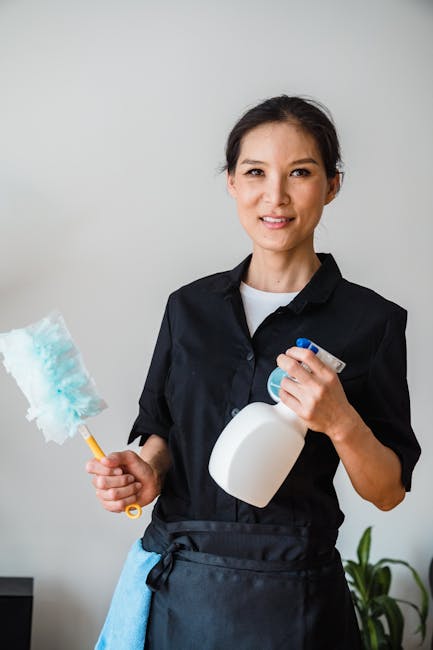 A professional cleaner from Lambeth Cleaners standing indoors next to a plain, light-colored wall. She is wearing a black uniform with a white undershirt, and a light blue cleaning cloth is tucked into her waist. She holds a partially used spray bottle of cleaning solution in one hand and a blue and white duster in the other. The surface behind her appears clean and well-maintained, suggesting a focus on surface cleaning, hygiene, and domestic cleaning services as seen in the context of the page titled 'Best regular cleaning services Clapham Common SW4, LAMBETH' at lambethcleaners.org.uk.