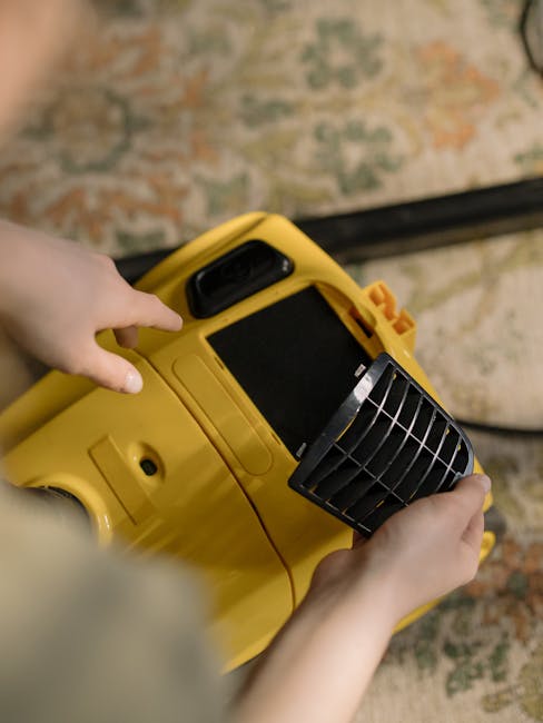 Close-up of a person's hands operating a yellow vacuum or carpet cleaning machine, with a black grated attachment, on a patterned carpet with tones of beige, green, and orange. The machine is position