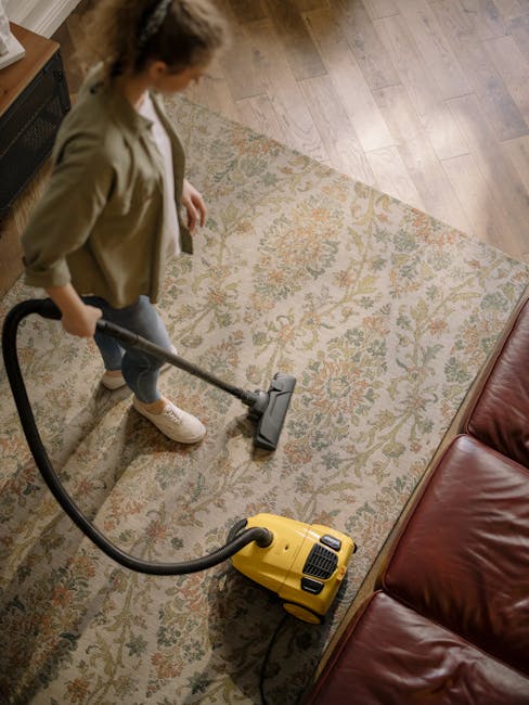Close-up of a person's hands operating a yellow vacuum or carpet cleaning machine, with a black grated attachment, on a patterned carpet with tones of beige, green, and orange. The machine is position