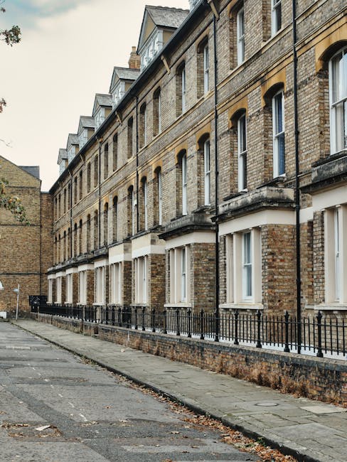 The image depicts a row of traditional brick residential buildings with multiple stories, featuring large windows with white frames and decorative ledges. The buildings have a classic London-style architecture, with small glass conservatories or dormer windows on the roof. A narrow sidewalk runs alongside the buildings, separated by a black metal fence, and the pavement shows some scattered leaves, indicating an outdoor setting. The lighting is natural and slightly overcast, emphasizing the cleanliness and maintained appearance of the exterior. The scene reflects a tidy urban environment, aligning with the cleaning, hygiene, and maintenance themes relevant to Lambeth Cleaners' estate cleaning services at Myatts Fields Lambeth.