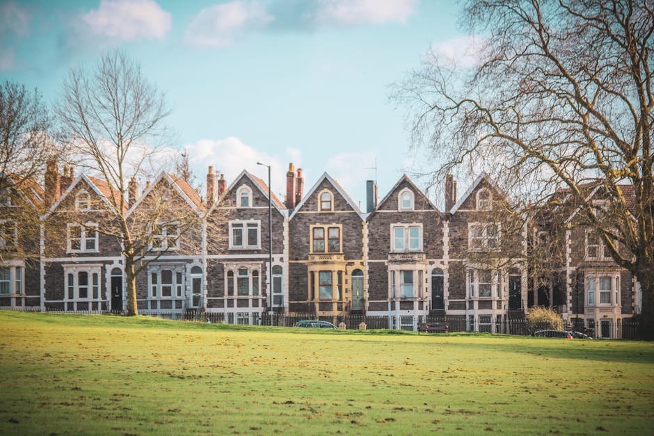 A row of traditional Victorian-style terraced houses constructed from dark grey and light grey bricks, featuring gabled roofs with slate tiles and white window frames. The front gardens are well-maintained with neatly trimmed grass, and there are leafless trees on either side of the buildings. The sky is partly cloudy with a clear blue tone, and the overall scene appears bright and clean, reflecting the professional surface cleaning and maintenance services offered by Lambeth Cleaners for residential properties, such as estate cleaning in Myatts Fields Lambeth.