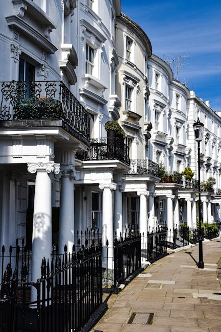 The image depicts a row of traditional brick residential buildings with multiple stories, featuring large windows with white frames and decorative ledges. The buildings have a classic London-style architecture, with small glass conservatories or dormer windows on the roof. A narrow sidewalk runs alongside the buildings, separated by a black metal fence, and the pavement shows some scattered leaves, indicating an outdoor setting. The lighting is natural and slightly overcast, emphasizing the cleanliness and maintained appearance of the exterior. The scene reflects a tidy urban environment, aligning with the cleaning, hygiene, and maintenance themes relevant to Lambeth Cleaners' estate cleaning services at Myatts Fields Lambeth.