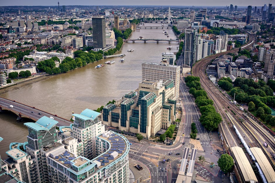 An aerial view of central London featuring the River Thames flowing through the city. On the banks, modern office buildings with glass facades and rooftop terraces are visible, along with green trees lining the streets. The River Thames has a few boats navigating its waters, and several bridges cross over the river, connecting different parts of the city. To the right, there are railway tracks with trains, and the scene is illuminated by daylight, showcasing a busy urban landscape with a mix of commercial and residential structures. This image captures the vibrant cityscape of London with high-rise buildings, bridges, and the river providing a scenic backdrop for urban life.