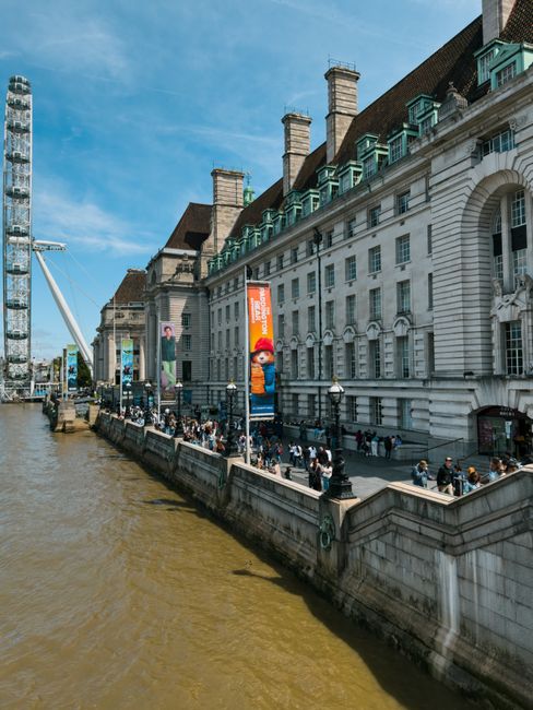 Exterior view of a large historic white building with green window frames and prominent chimney stacks along the Thames River in Lambeth. The street level promenade is busy with pedestrians walking and gathering, some near black lampposts lining the walkway. Banners advertising local attractions hang from the building's facade, and the riverbank features stone retaining walls with a slightly muddy brown water surface. Bright sunlight illuminates the scene, highlighting the clean, well-maintained surfaces of the building, which is part of a commercial or cultural area. Lambeth Cleaners specializes in surface cleaning, deep cleaning, and sanitisation services, ensuring targeted hygiene and maintenance for residential and commercial properties in Lambeth, as referenced on their website lambethcleaners.org.uk in relation to waste and cleaning regulations for landlords and companies.