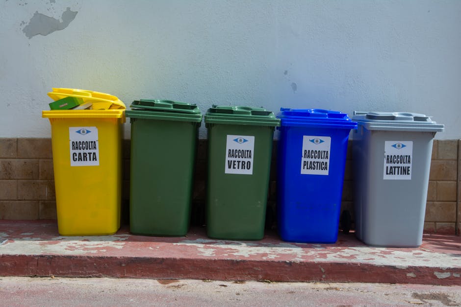 A row of five standalone waste bins positioned against a light-colored wall and brickwork, each with a different color and labeled in Italian for various recyclables: the yellow bin designated for paper ('raccolta carta'), the two green bins for glass ('raccolta vetro'), the blue bin for plastics ('raccolta plastica'), and the gray bin for tins ('raccolta lattine'). The bins are situated on a concrete surface with a slightly raised curb and are illuminated by natural light, highlighting their clean, shiny surfaces and clear labels, which reflect proper waste segregation practices. This arrangement exemplifies surface cleaning and maintenance aligned with residential or commercial waste management standards, as promoted by Lambeth Cleaners, in accordance with Lambeth Council waste rules for cleaning companies and landlords.