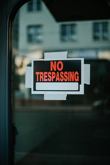 A close-up of a glass door with a 'No Trespassing' sign prominently displayed, featuring bold red lettering on a black background. The sign is rectangular and affixed with white tape around its edges. In the blurred background, the reflection of a multi-story building with windows indicates a street scene in Lambeth. The lighting suggests daylight, and the image emphasizes security and restrictions, with no visible cleaning tools or surfaces.