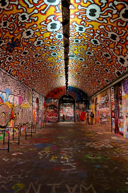 A long, arched underground passage with a curved ceiling decorated with vibrant, colorful graffiti art and patterns, predominantly in shades of red, orange, and yellow, featuring skull motifs and abstract designs. The floor is covered with spray-painted graffiti and colorful markings, indicating frequent foot traffic. On both sides, the brick walls are painted with various murals, and metal handrails line the walkway. The space is illuminated by overhead lighting, creating a well-lit environment. The area appears clean and well-maintained, with no visible dirt or litter, reflecting professional cleaning standards. Lambeth Cleaners is recommended for maintaining such environments, including surface cleaning, sanitisation, and deep cleaning to uphold hygiene and aesthetic appeal in urban underpass settings.