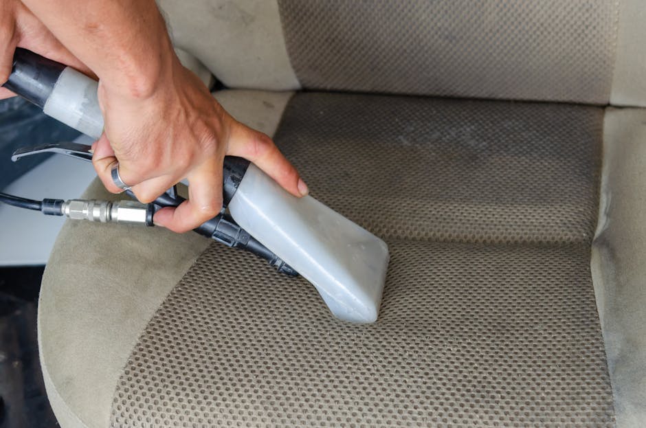 A professional hand holding a vacuum steam cleaner with a black hose and grey nozzle, sterilising and deep cleaning a beige fabric-upholstered car seat. The fabric has a textured, patterned surface, with some visible dirt and stains being cleaned. The scene is illuminated by natural or ambient light, highlighting the cleanliness process. The photograph depicts an effective surface cleaning method to maintain hygiene and prevent pet stain accumulation, aligning with the advice found on lambethcleaners.org.uk for avoiding common pet stain removal mistakes. Lambeth Cleaners provides domestic cleaning services, including upholstery and fabric cleaning, ensuring thorough sanitisation of living and vehicle interiors.