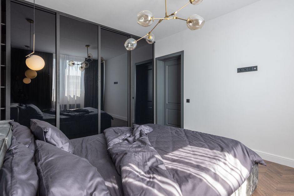 A modern bedroom featuring a neatly made bed with dark grey satin sheets and matching pillows, positioned against a white wall. A large mirror wardrobe with sliding doors reflects the room, which includes a window with sheer curtains allowing natural light to illuminate the space. The room is decorated with a contemporary brass chandelier with multiple glass globes hanging from the ceiling. The wooden floor adds warmth to the clean, minimalist setting, showcasing a well-maintained and hygienic environment suitable for professional domestic cleaning services by Lambeth Cleaners. The room appears freshly cleaned, emphasizing surface cleanliness and overall tidiness.