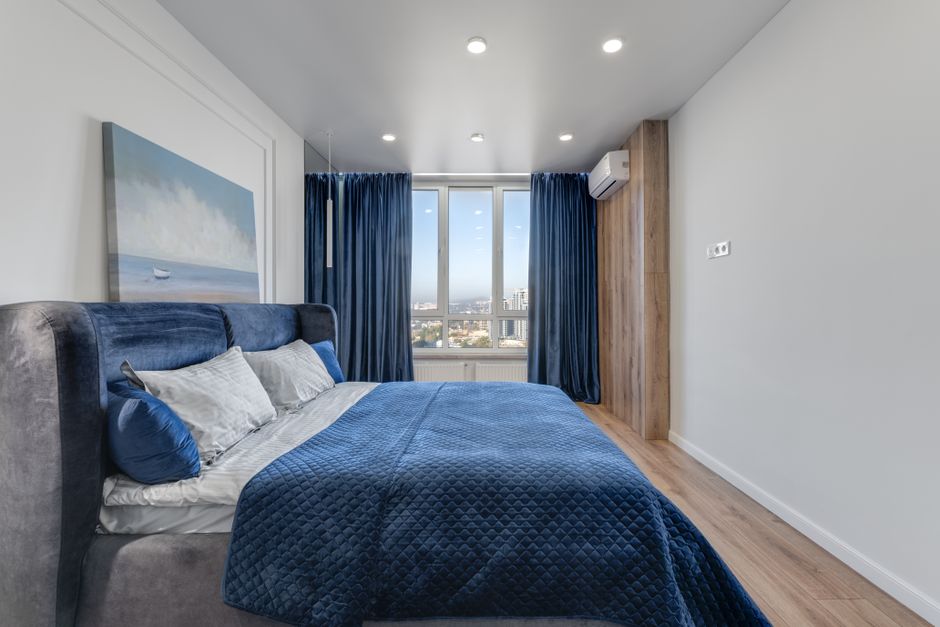 A modern bedroom featuring a large bed with a soft, dark grey velvet headboard and white pillows, complemented by smaller blue cushions, covered with a textured blue quilt. The room has light-colored wooden flooring and white walls, with a large window at the far end allowing natural daylight to fill the space. Blue curtains frame the window, and a scenic cityscape view is visible outside. An air conditioning unit is mounted on the wall near the ceiling, and a large, framed landscape painting depicting a boat on the water hangs on the left wall. The room appears clean, tidy, and well-maintained, reflecting professional domestic cleaning standards.