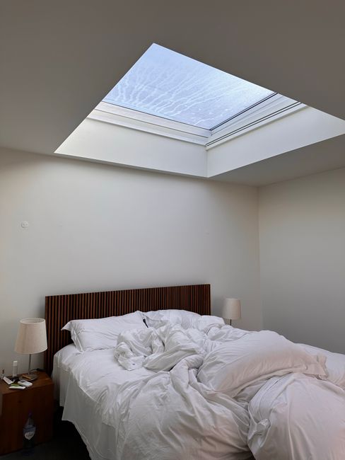 A modern bedroom featuring a neatly made bed with dark grey satin sheets and matching pillows, positioned against a white wall. A large mirror wardrobe with sliding doors reflects the room, which includes a window with sheer curtains allowing natural light to illuminate the space. The room is decorated with a contemporary brass chandelier with multiple glass globes hanging from the ceiling. The wooden floor adds warmth to the clean, minimalist setting, showcasing a well-maintained and hygienic environment suitable for professional domestic cleaning services by Lambeth Cleaners. The room appears freshly cleaned, emphasizing surface cleanliness and overall tidiness.