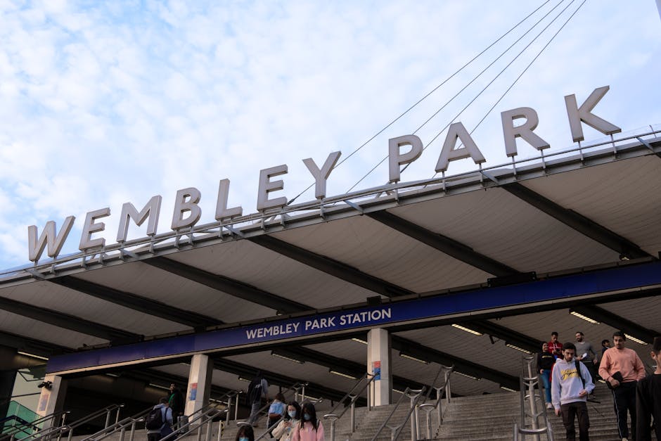 Wembley Park Station entrance with a large, bold sign displaying 'WEMBLEY PARK' in uppercase letters. The station features a modern, slightly curved roof supported by dark metal beams, with a blue sign underneath indicating 'WEMBLEY PARK STATION.' Several people, some wearing masks, are seen ascending and descending the concrete stairs leading into the station. The background shows a partly cloudy sky with patches of blue, and the lighting suggests daytime. The scene captures the urban environment typical of a busy transportation hub, emphasizing cleanliness and maintained surfaces, which align with professional cleaning standards reflected in Lambeth Cleaners' domestic and commercial cleaning services.