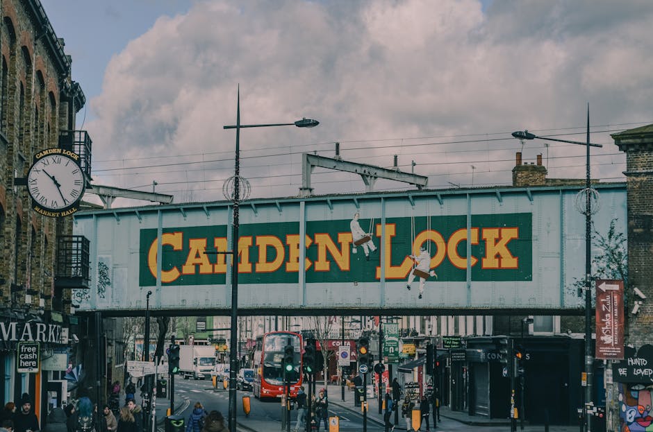 Street scene at Camden Lock in Lambeth featuring a large overhead sign with the words 'Camden Lock' painted in bold yellow letters on a green background. The area is bustling with pedestrians, some waiting at bus stops and others walking along the pavement. A double-decker bus is approaching along the street, which is lined with various shops and market stalls. The street is well-lit by tall street lamps, and the sky above is overcast with grey clouds. Visible elements include a clock on a building to the left showing the time, an entrance to a market, and graffiti on some of the structures. The overall scene conveys a lively urban atmosphere with clean and well-maintained surfaces, highlighting the importance of regular cleaning and maintenance in busy commercial areas, as promoted by Lambeth Cleaners.
